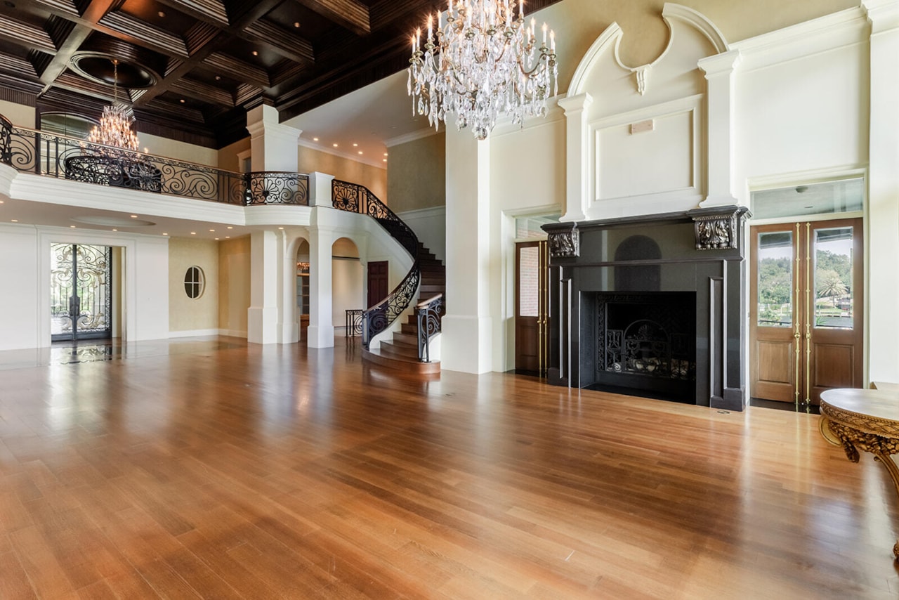 The two-story great room with coffered dark wood ceiling, twin crystal chandeliers, a curved iron-railed staircase, and a black marble fireplace surround