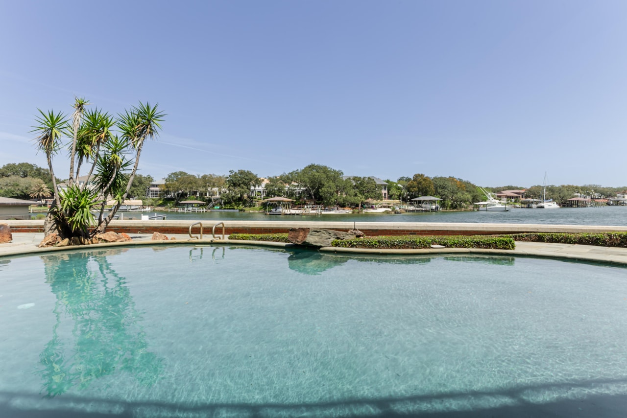 An infinity-edge pool with a stone deck facing the protected cove, palms in raised planters and boats moored across the water