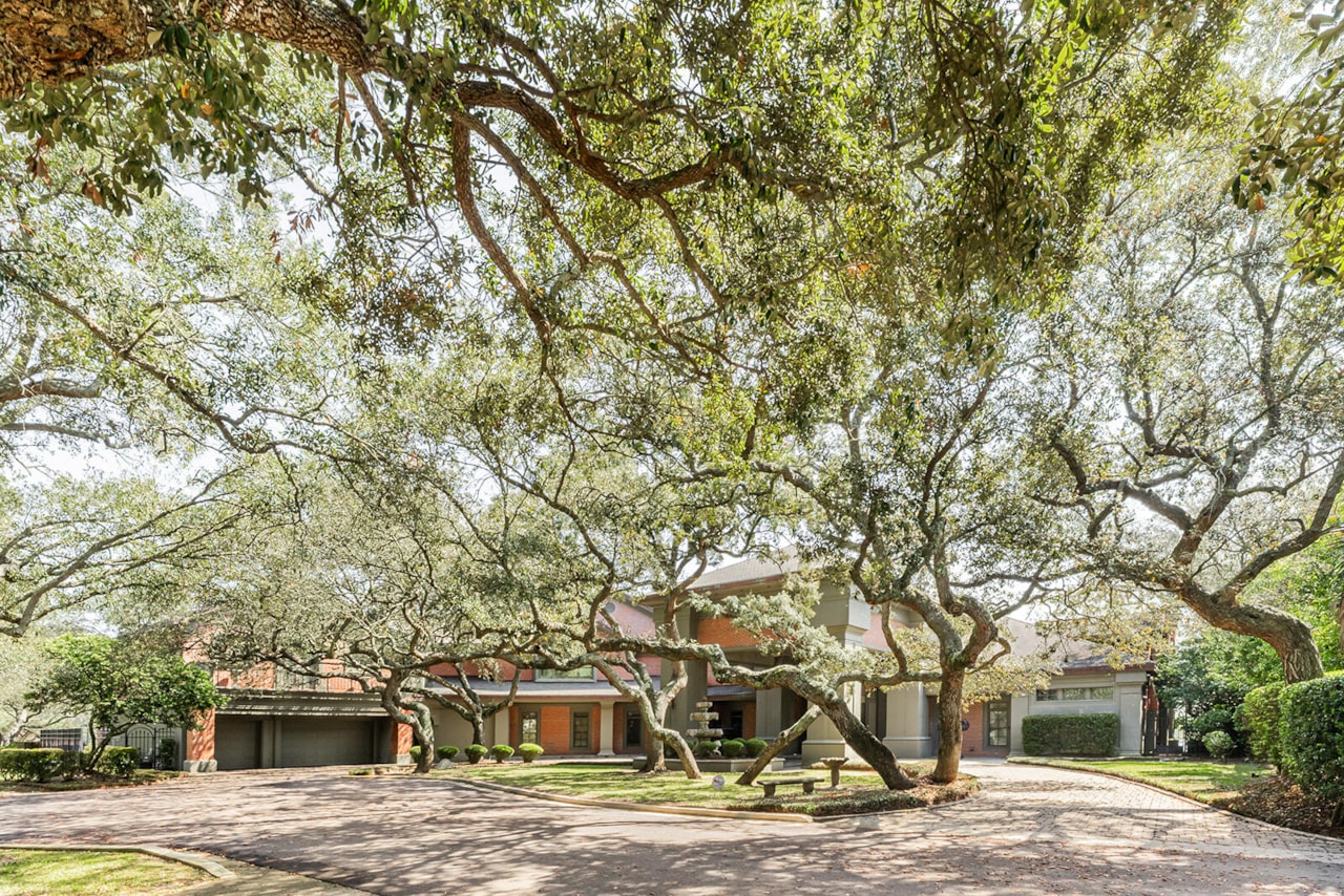 The front elevation of the brick villa seen through a canopy of mature live oaks, with a circular cobble courtyard and tiered fountain